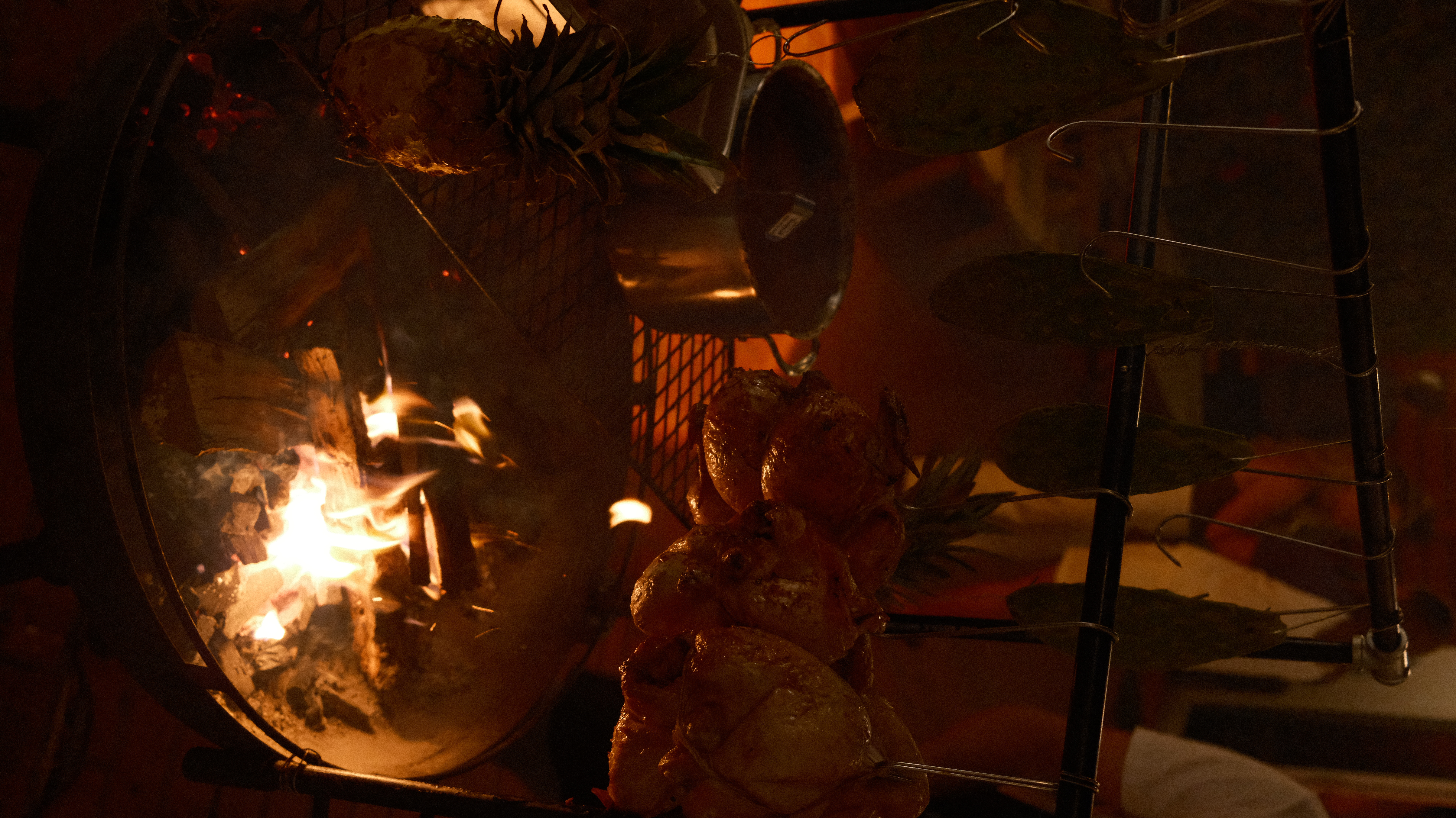 Whole chickens, pineapple, and vegetables hanging over an open fire at a Motxo event