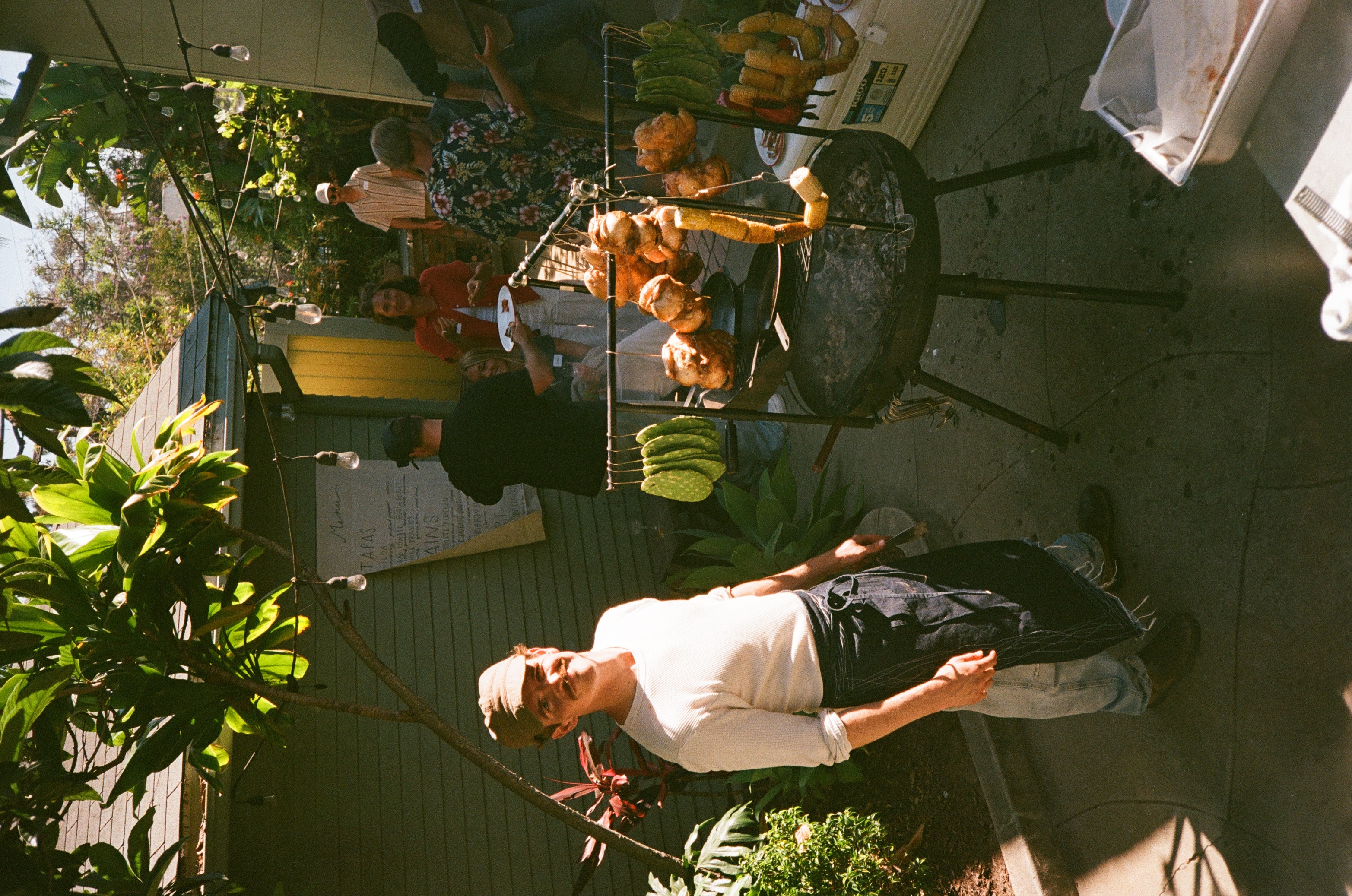 Zack beside the backyard live-fire rig with hanging chickens, nopales, peppers, and corn